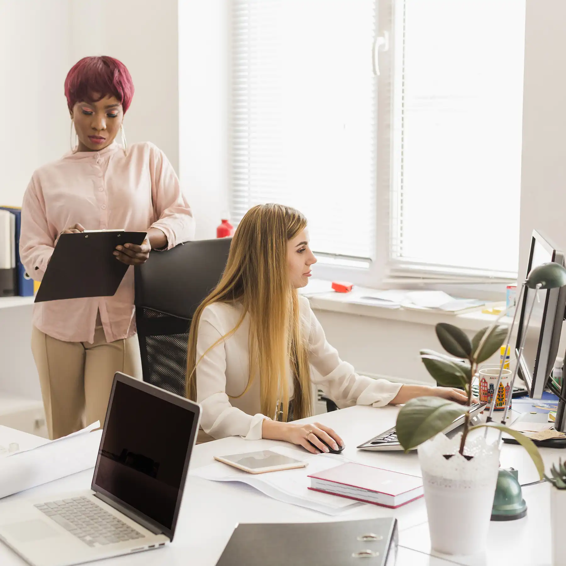 A colleague with a clipboard checks in with a team member working at her desk